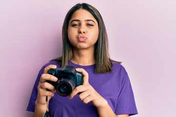 Young latin woman using reflex camera puffing cheeks with funny face. mouth inflated with air,...