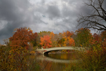 Bow Bridge is reflected in the water of Central Park in NYC, surrounded by brilliant fall foliage, under a stormy sky.