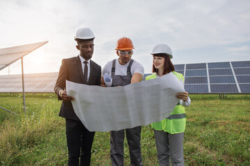 Two engineers and one technician examining drawings of solar farm while standing together outdoors. Male and female partners in helmets working on modern business project.