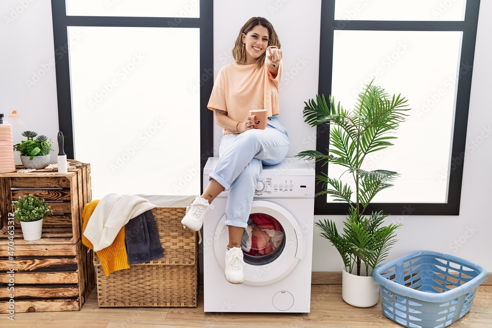 Sticker young hispanic woman drinking coffee waiting for washing machine at laundry room pointing to you and