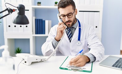Fototapeta premium Young hispanic man wearing doctor uniform talking on the telephone and write on clipboard at clinic