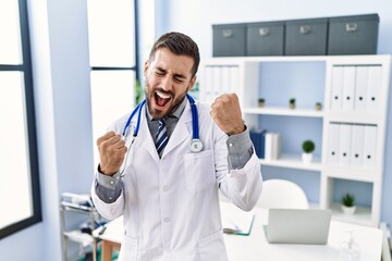 Naklejka na ściany i meble Handsome hispanic man wearing doctor uniform and stethoscope at medical clinic very happy and excited doing winner gesture with arms raised, smiling and screaming for success. celebration concept.