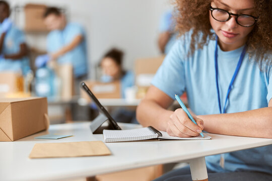 Cropped shot of young female volunteer in blue uniform making notes, using tablet pc while working on donation project. Team sorting, packing items in the background