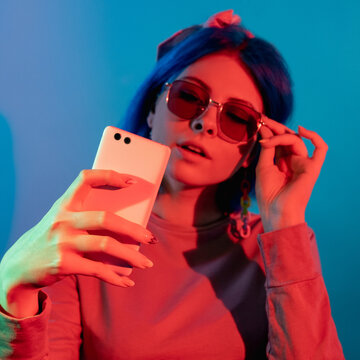 Phone selfie. Gadget people. Cyber technology. Stylish millennial woman taking photo on smartphone red neon light isolated on blue background.
