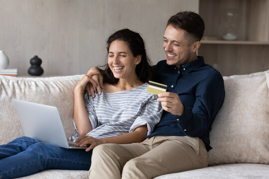 Young millennial Latin couple sit on sofa use laptop holding credit card buying on internet goods for home. Happy online retail web services clients, on-line purchase, instant money transfer concept