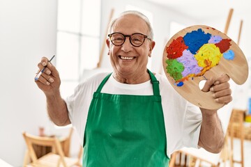 Senior grey-haired artist man smiling happy holding paintbrush and palette at art studio.