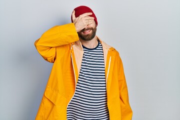 Caucasian man with beard wearing yellow raincoat smiling and laughing with hand on face covering eyes for surprise. blind concept.
