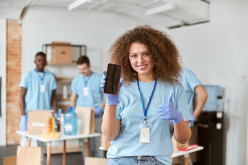 Young woman, volunteer in blue uniform holding, showing smartphone to camera and smiling while standing indoors. Team sorting, packing items in the background