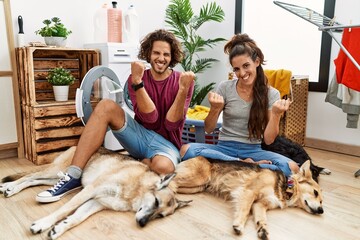 Young hispanic couple doing laundry with dogs very happy and excited doing winner gesture with arms raised, smiling and screaming for success. celebration concept.