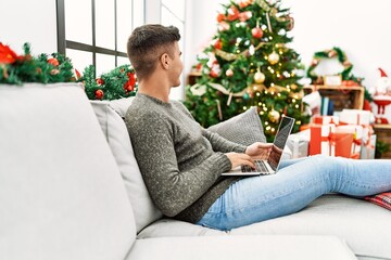 Young hispanic man using laptop sitting on sofa by christmas tree at home