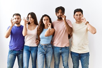 Group of young people standing together over isolated background mouth and lips shut as zip with fingers. secret and silent, taboo talking