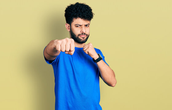 Young arab man with beard wearing casual blue t shirt punching fist to fight, aggressive and angry attack, threat and violence