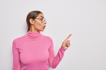 Horizontal shot of shocked surprised woman reacts on amazing news wears transparent glassed and casual pink turtleneck points at blank copy space advertises something isolated over white background