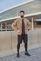 Full length shot of motivated bearded sportsman keeps hands on waist dressed in active wear...