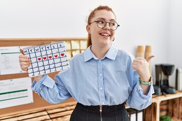 Young redhead woman holding heart calendar at the office pointing thumb up to the side smiling happy with open mouth
