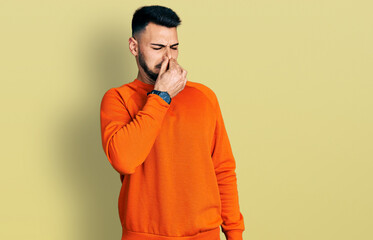 Young hispanic man with beard wearing casual orange sweater smelling something stinky and disgusting, intolerable smell, holding breath with fingers on nose. bad smell