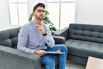 Young psychologist man at consultation office thinking concentrated about doubt with finger on chin and looking up wondering