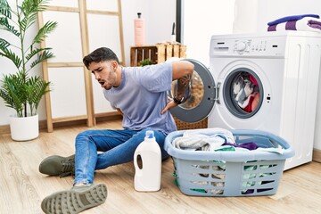 Young hispanic man putting dirty laundry into washing machine suffering of backache, touching back with hand, muscular pain