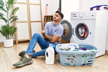 Young hispanic man putting dirty laundry into washing machine looking to the side with arms crossed...