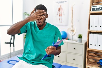 Young african american man working at pain recovery clinic peeking in shock covering face and eyes with hand, looking through fingers with embarrassed expression.