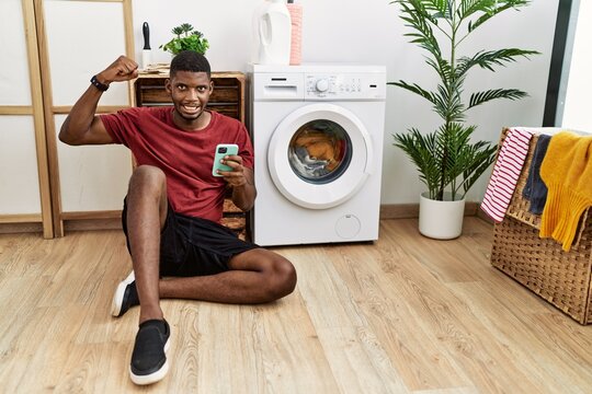 Young african american man using smartphone waiting for washing machine strong person showing arm muscle, confident and proud of power