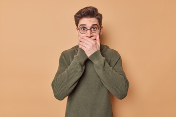 Fototapeta premium Emotive scared man tries to be speechless covers mouth with hands checks out big new wears round spectacles and jumper poses against brown background witnesses disaster frightened to make sound