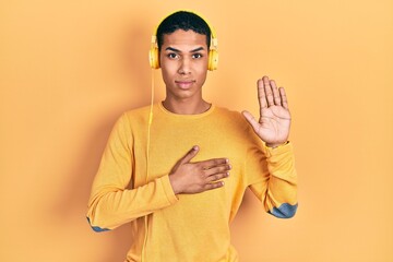Young african american guy listening to music using headphones swearing with hand on chest and open palm, making a loyalty promise oath