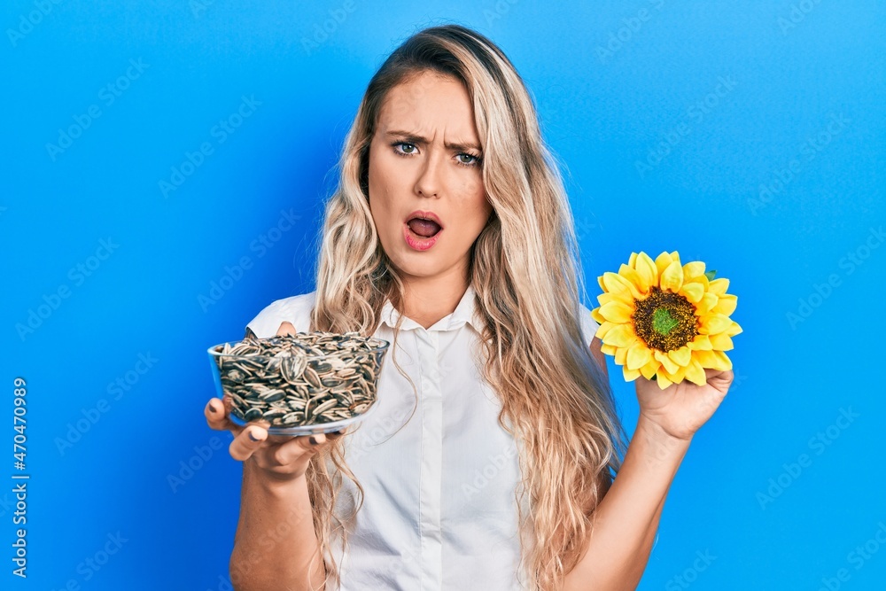 Sticker beautiful young blonde woman holding sunflower seeds an flower in shock face, looking skeptical and 