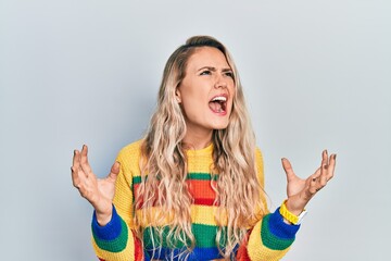 Beautiful young blonde woman wearing colored sweater crazy and mad shouting and yelling with aggressive expression and arms raised. frustration concept.