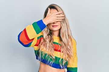 Beautiful young blonde woman wearing colored sweater covering eyes with hand, looking serious and sad. sightless, hiding and rejection concept