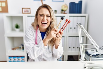 Young beautiful doctor woman holding diapason instrument smiling and laughing hard out loud because funny crazy joke.