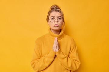 Fototapeta premium Coy beautiful woman purses lips and asks for apology keeps palms pressed together needs something purses lips has sad expression wears optical glasses and sweater isolated on yellow studio wall