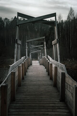Obraz premium Old and gloomy wooden bridge over the river. Detachment in the water. Gloomy and mysterious forest. Cloudy sky. Wooden structure of an old abandoned bridge.