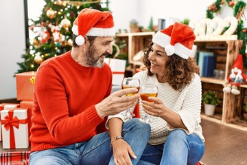Middle age hispanic couple wearing christmas hat. Sitting on the floor toasting with champagne at home.