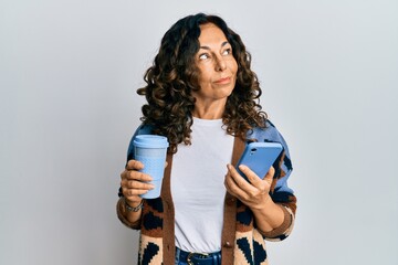 Middle age hispanic woman drinking a cup of coffee and looking at the smartphone screen smiling looking to the side and staring away thinking.