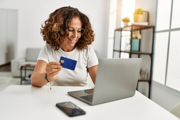 Middle age hispanic woman using laptop holding credit card at home