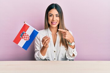 Young hispanic woman holding croatia flag sitting on the table smiling happy pointing with hand and finger - obrazy, fototapety, plakaty