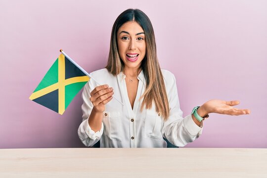 Young hispanic woman holding jamaica flag sitting on the table celebrating achievement with happy smile and winner expression with raised hand