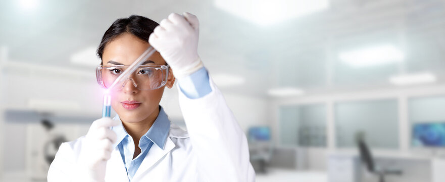 Young focused female chemist in lab coat, gloves and protective eyeglass dropping a blue substance into a test tube with a pipette in laboratory. Chemical, pharmaceutical or medical research concept.