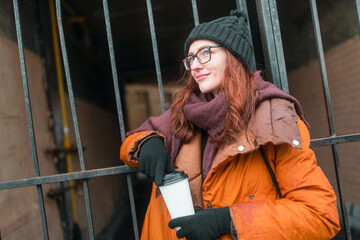 Fototapeta na wymiar Woman drinks coffee in the cold winter. Warm up in weather with a hot drink. Warm orange jacket and purple scarf. In the background is a large iron tails.