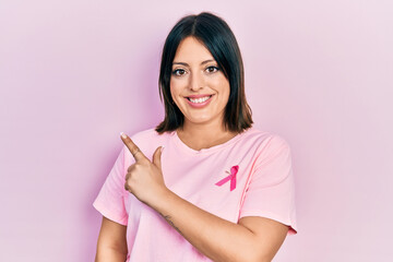 Young hispanic woman wearing pink cancer ribbon on t shirt cheerful with a smile on face pointing...