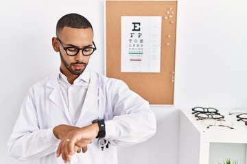 African american optician man standing by eyesight test checking the time on wrist watch, relaxed...