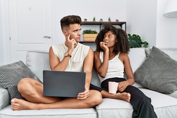 Obraz na płótnie Canvas Young interracial couple using laptop at home sitting on the sofa looking stressed and nervous with hands on mouth biting nails. anxiety problem.