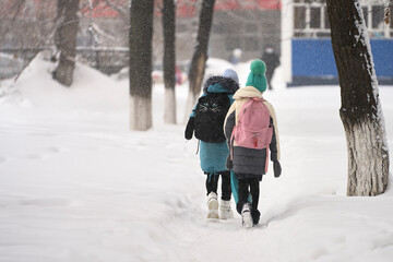 Winter city landscape. two schoolgirls go to school on a snow-covered trail. Snow is falling. Selective focus. - obrazy, fototapety, plakaty