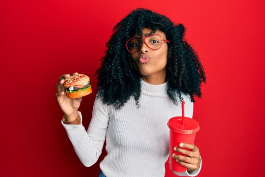 African american woman with afro hair eating a tasty classic burger and soda looking at the camera blowing a kiss being lovely and sexy. love expression.