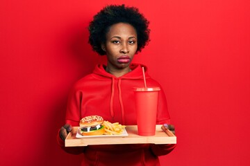 Young african american woman eating a tasty classic burger with fries and soda skeptic and nervous, frowning upset because of problem. negative person.