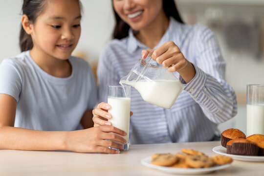 Asian Mother Pouring Fresh Milk For Her Cute Teen Daughter, Pretty Girl Having Breakfast With Mom, Sitting In Kitchen