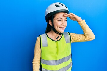 Beautiful brunette little girl wearing bike helmet and reflective vest very happy and smiling looking far away with hand over head. searching concept.