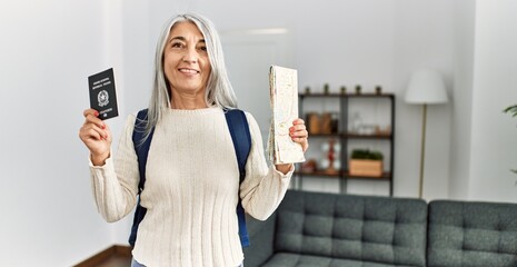 Middle age grey-haired woman wearing backpack holding map and passport at home