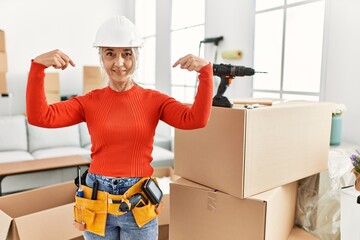 Middle age grey-haired woman wearing hardhat standing at new home looking confident with smile on face, pointing oneself with fingers proud and happy.